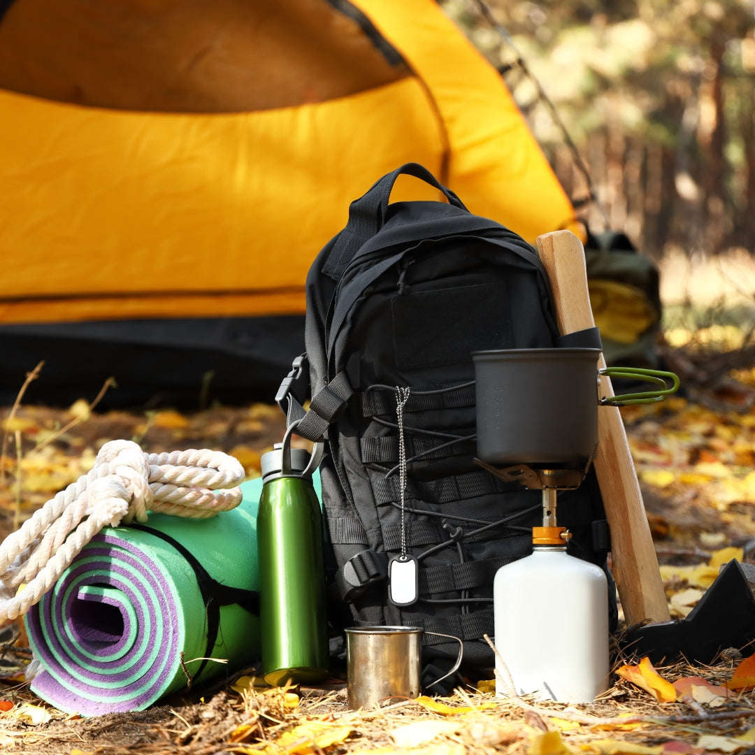 A black camping backpack stands on a leaf-covered forest floor with a yellow tent blurred in the background. Surrounding the backpack are camping items including a rolled green and purple sleeping mat, a green water bottle, a metal cup, a white fuel canister, a wooden spoon, and a rope.