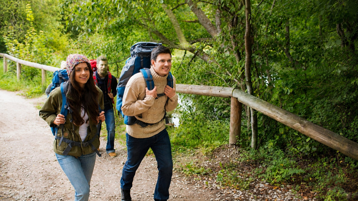 Three people hiking with large backpacks on a dirt path surrounded by green trees and a wooden fence.