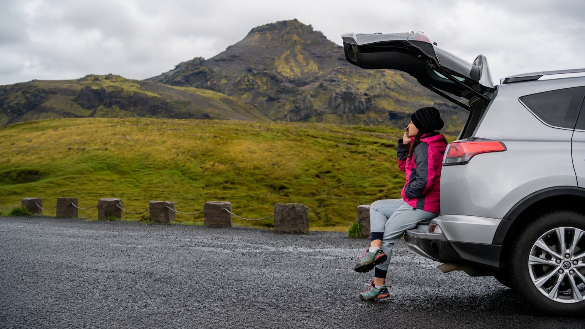 A woman in a pink jacket and gray pants sits on the open trunk of a silver SUV on a gravel road, with a dramatic green and brown mountain landscape in the background under an overcast sky.