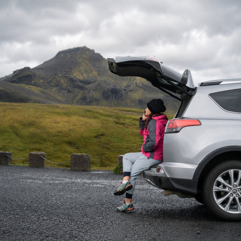 A woman in a pink jacket and gray pants sits on the open trunk of a silver SUV on a gravel road, with a dramatic green and brown mountain landscape in the background under an overcast sky.